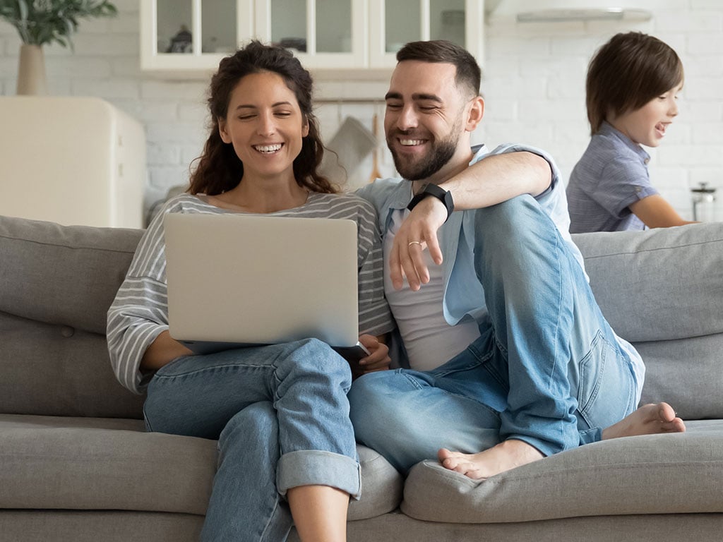 Parents on couch looking at laptop with child running in the background