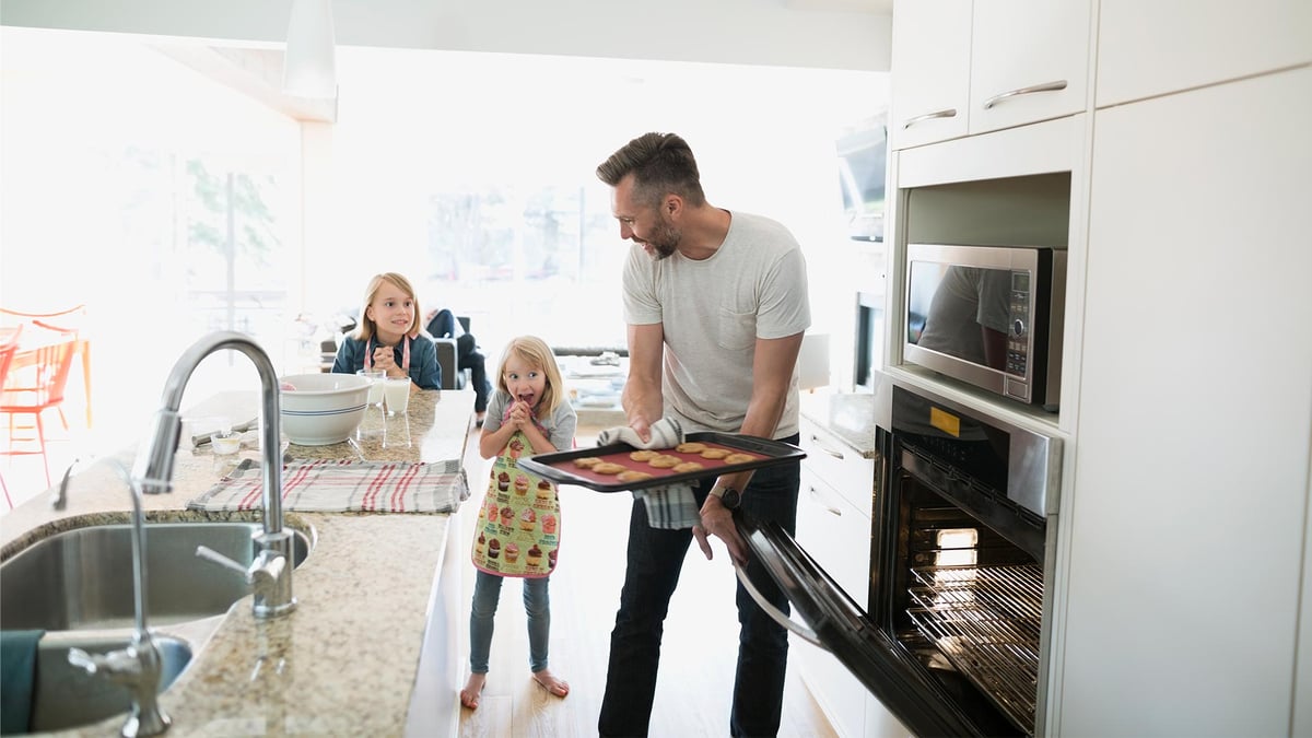 Father pulling out baked cookies out of the oven while his two young daughters look on excitedly