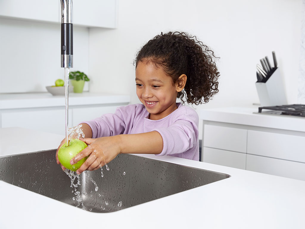 Little girl washing green apple at kitchen sink.